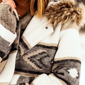 May include: A cream-colored jacket with a geometric pattern in black, tan, and gray. The jacket features a faux fur-lined hood and a button closure. The sleeves have a striped design, and the overall style is a cozy, Southwestern-inspired look.
