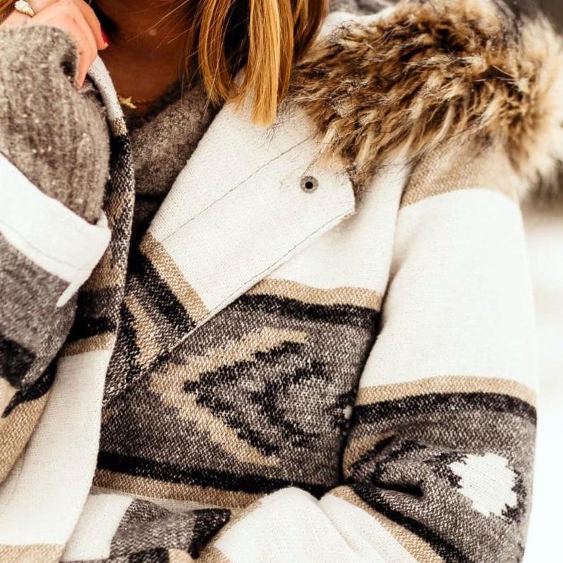 May include: A cream-colored jacket with a geometric pattern in black, tan, and gray. The jacket features a faux fur-lined hood and a button closure. The sleeves have a striped design, and the overall style is a cozy, Southwestern-inspired look.