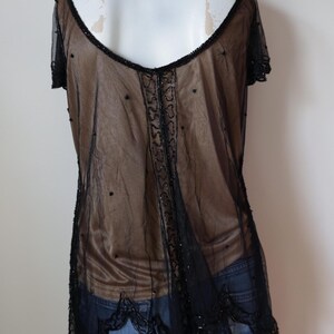 May include: A sheer black top with short sleeves and a low back. The top is embellished with black beads along the neckline, center, and hem. The garment is layered over a brown fabric, creating a contrast.