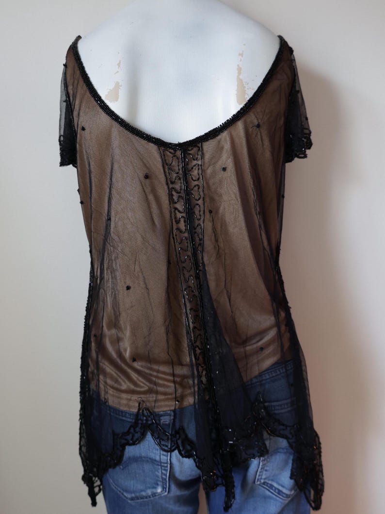 May include: A sheer black top with short sleeves and a low back. The top is embellished with black beads along the neckline, center, and hem. The garment is layered over a brown fabric, creating a contrast.