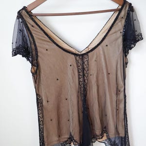 May include: A vintage sheer top with a V-neck design, hanging on a wooden hanger. The top features a nude-colored underlay with a black overlay adorned with beaded accents and a scalloped lace trim. The short sleeves are also black.