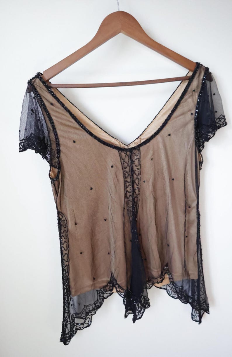 May include: A vintage sheer top with a V-neck design, hanging on a wooden hanger. The top features a nude-colored underlay with a black overlay adorned with beaded accents and a scalloped lace trim. The short sleeves are also black.
