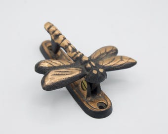 Puede incluir: Un aldaba de puerta con forma de libélula de color bronce. La libélula tiene alas y cuerpo detallados, montada sobre una base rectangular con orificios para tornillos. La aldaba es de metal y tiene un acabado envejecido.