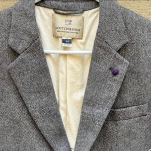 Op de afbeelding: Een grijze tweed blazer met een visgraatpatroon. De blazer heeft een gekerfde revers, een paarse reversspeld en een zak. De binnenvoering is crèmekleurig en het label luidt "SCOTCH & SODA AMSTERDAM COUTURE."