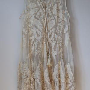 Puede incluir: Vestido sin mangas color marfil con intrincados bordados en forma de hojas. El vestido presenta un escote en V, un lazo con borlas y una capa transparente. Cuelga de una percha blanca.