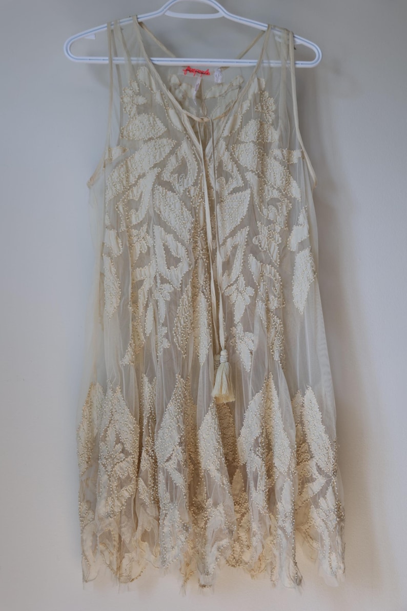 Puede incluir: Vestido sin mangas color marfil con intrincados bordados en forma de hojas. El vestido presenta un escote en V, un lazo con borlas y una capa transparente. Cuelga de una percha blanca.