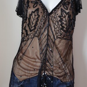 May include: Black beaded top with a V-neck and short sleeves. The sheer overlay features intricate leaf and geometric patterns. The top is worn over a nude-colored underlayer and paired with blue jeans.
