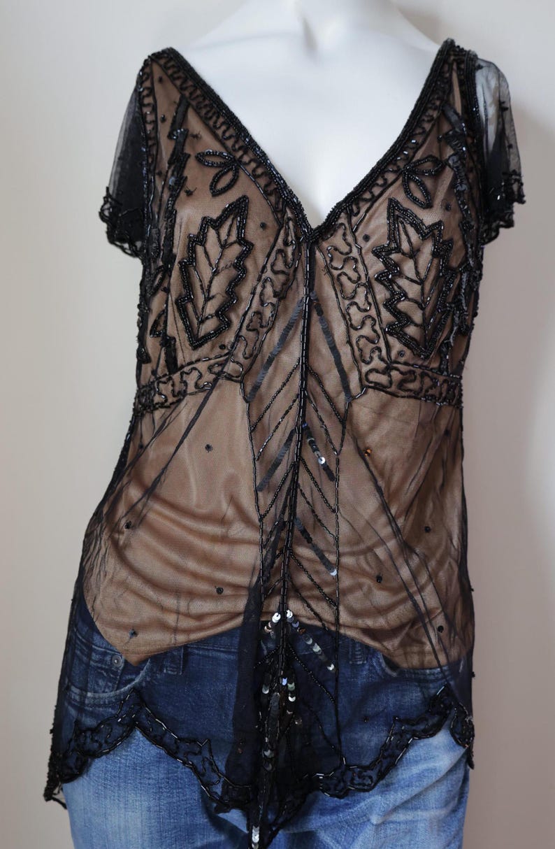 May include: Black beaded top with a V-neck and short sleeves. The sheer overlay features intricate leaf and geometric patterns. The top is worn over a nude-colored underlayer and paired with blue jeans.