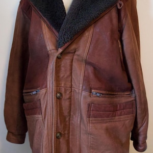 Peut inclure: Veste en cuir marron avec un col en peau de mouton gris foncé. La veste a une fermeture boutonnée, deux poches zippées et deux poches à rabat. La veste est présentée sur un mannequin.