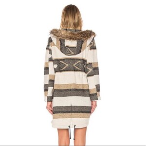 May include: A long coat with a hood and faux fur trim. The coat features a striped pattern in shades of beige, brown, and gray, with a geometric design on the upper back. It has a drawstring waist and a zipper closure.