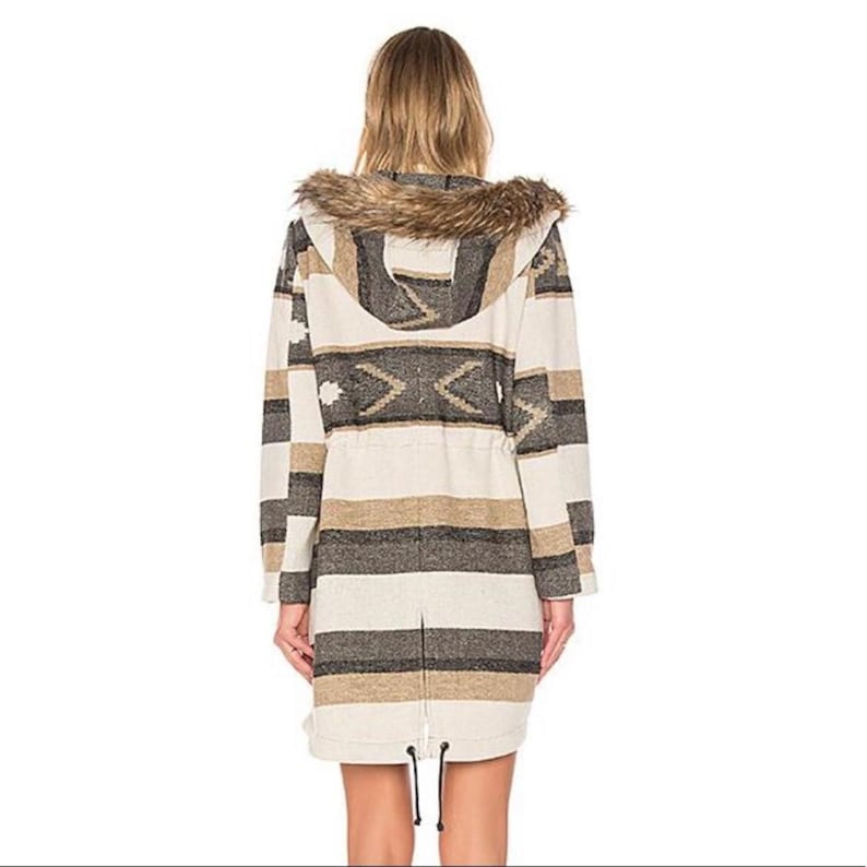 May include: A long coat with a hood and faux fur trim. The coat features a striped pattern in shades of beige, brown, and gray, with a geometric design on the upper back. It has a drawstring waist and a zipper closure.