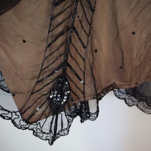 May include: Close-up of a black, sheer garment with intricate bead and sequin embellishments. The design features a central, stylized feather motif with a scalloped lace trim along the edge. The fabric appears to be layered over a tan underlay.