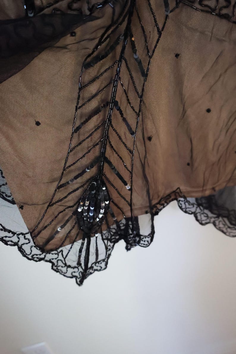 May include: Close-up of a black, sheer garment with intricate bead and sequin embellishments. The design features a central, stylized feather motif with a scalloped lace trim along the edge. The fabric appears to be layered over a tan underlay.