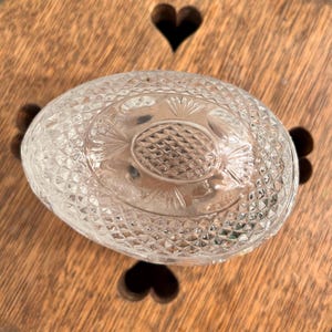 Op de afbeelding: Een heldere, ovaalvormige glazen container met een diamantgeslepen patroon. De container heeft een gedetailleerd, gestructureerd ontwerp aan de bovenkant. Hij staat op een houten oppervlak met hartvormige uitsparingen.