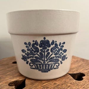 May include: Cream ceramic plant pot with a blue floral pattern. The design shows a basket of flowers with leaves and other floral elements. The pot has a wide rim and a slightly tapered body.