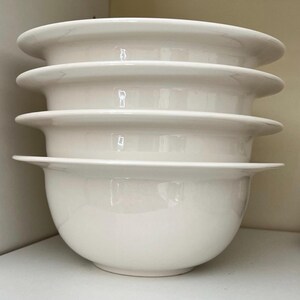 Op de afbeelding: Vier witte keramische kommen op elkaar gestapeld. Elke kom heeft een afgeronde bodem en een brede, platte rand. De kommen zijn schoon en lijken ongebruikt. De kommen zijn waarschijnlijk bedoeld om voedsel in te serveren.