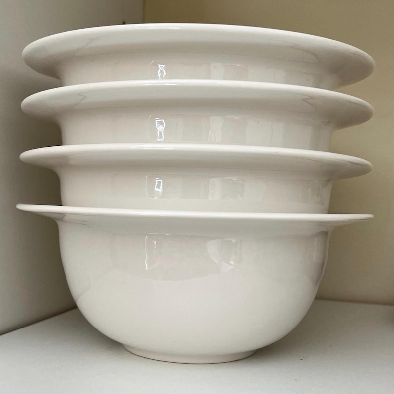 Op de afbeelding: Vier witte keramische kommen op elkaar gestapeld. Elke kom heeft een afgeronde bodem en een brede, platte rand. De kommen zijn schoon en lijken ongebruikt. De kommen zijn waarschijnlijk bedoeld om voedsel in te serveren.