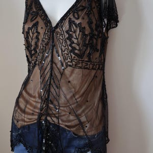 May include: A black beaded top with a deep V-neck and leaf-like patterns. The sheer overlay reveals a tan underlay, and the top is adorned with sequins. The garment has short sleeves and is paired with blue jeans.