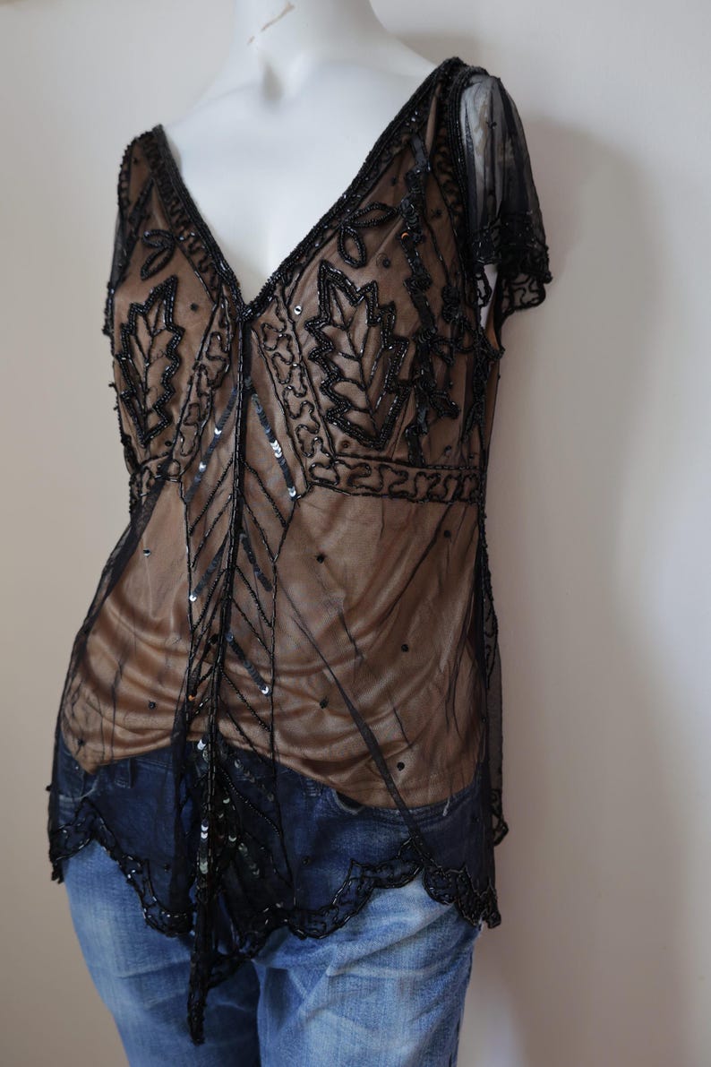 May include: A black beaded top with a deep V-neck and leaf-like patterns. The sheer overlay reveals a tan underlay, and the top is adorned with sequins. The garment has short sleeves and is paired with blue jeans.