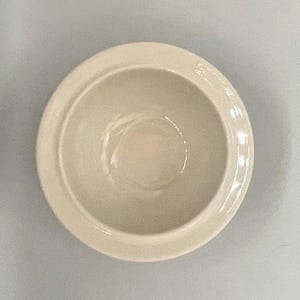 Op de afbeelding: Een cr&egrave;mekleurige keramische kom met een afgeronde vorm en een iets verhoogde rand. De kom heeft een gladde, glanzende afwerking en is leeg. De kom staat in het midden van het frame en lijkt nieuw.