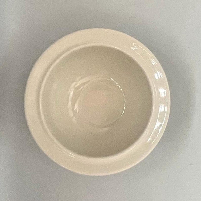 Op de afbeelding: Een cr&egrave;mekleurige keramische kom met een afgeronde vorm en een iets verhoogde rand. De kom heeft een gladde, glanzende afwerking en is leeg. De kom staat in het midden van het frame en lijkt nieuw.