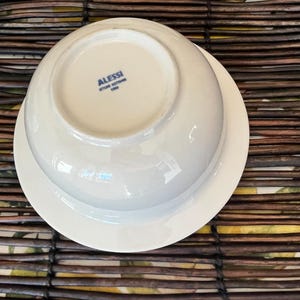 Op de afbeelding: Witte porseleinen kom- en schotelset. De kom is omgekeerd en onthult de merknaam "ALESSI" en het logo in blauw. De set staat op een bruin geweven oppervlak.