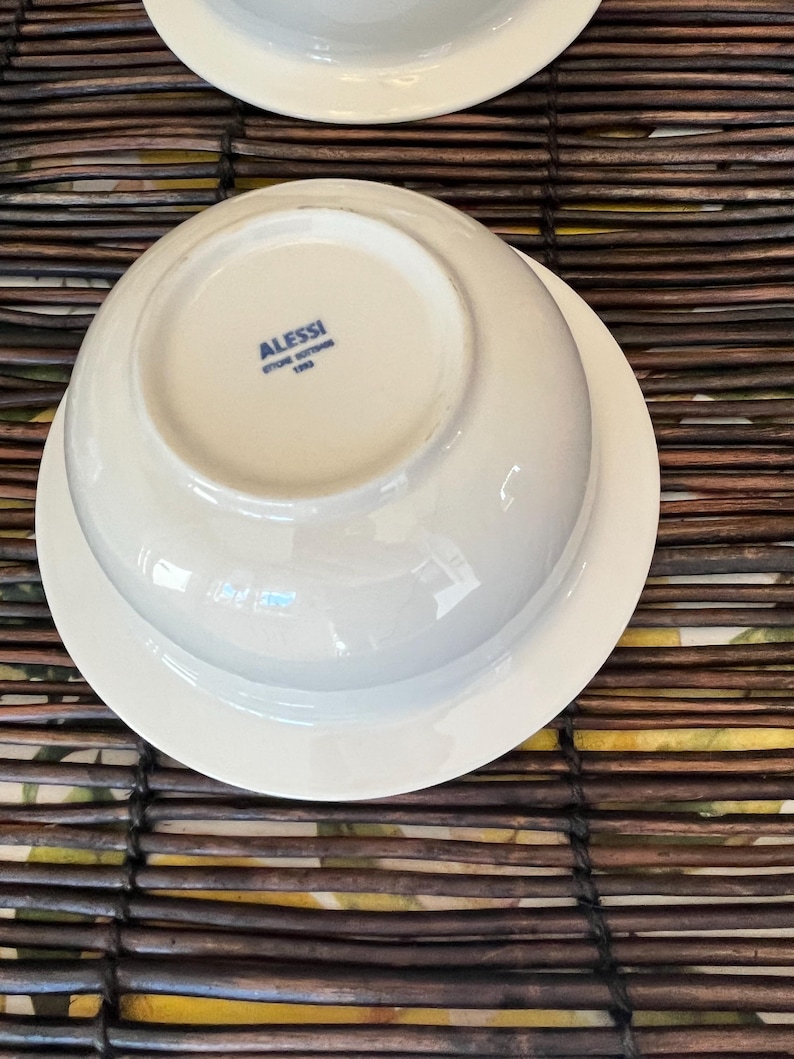 Op de afbeelding: Witte porseleinen kom- en schotelset. De kom is omgekeerd en onthult de merknaam "ALESSI" en het logo in blauw. De set staat op een bruin geweven oppervlak.