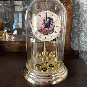 May include: A gold-tone anniversary clock with a glass dome, featuring a white dial with black Roman numerals and a central illustration of two figures in a landscape. The clock has a rotating pendulum with four gold spheres on a circular base.