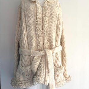 May include: An off-white, long-sleeved, zip-up cardigan featuring a chunky cable knit pattern, a textured bouclé trim along the collar, front zip, and hem, two front pockets, and a matching knit tie belt at the waist for a secure fit.