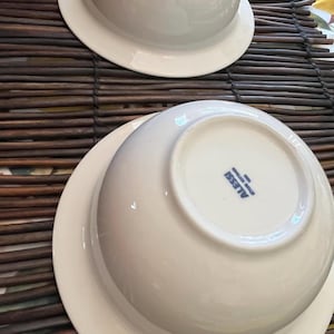 Op de afbeelding: Twee witte keramische kommen met een brede rand, gestapeld op een bruine geweven mat. De kommen staan ondersteboven en onthullen de onderkant met de merknaam "ALESSI" in blauw.