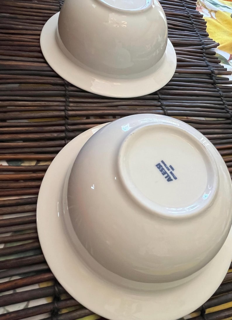 Op de afbeelding: Twee witte keramische kommen met een brede rand, gestapeld op een bruine geweven mat. De kommen staan ondersteboven en onthullen de onderkant met de merknaam "ALESSI" in blauw.