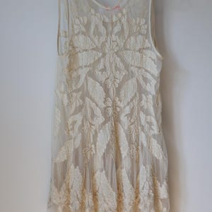 Puede incluir: Vestido sin mangas de color marfil con intrincados bordados florales. El vestido est&aacute; hecho de una tela transparente, con un patr&oacute;n detallado de hojas y flores. El vestido est&aacute; colgado en una percha blanca contra una pared blanca.