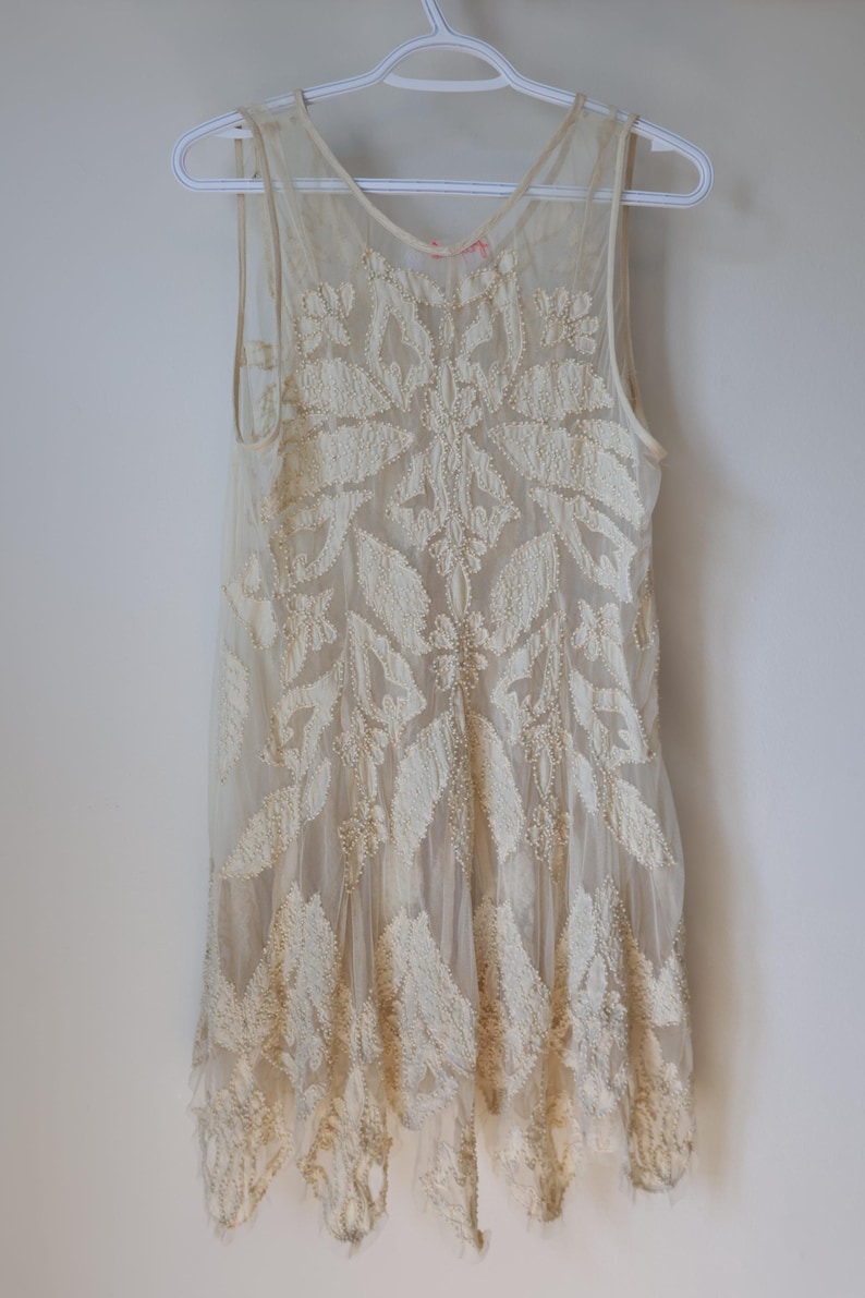 Puede incluir: Vestido sin mangas de color marfil con intrincados bordados florales. El vestido est&aacute; hecho de una tela transparente, con un patr&oacute;n detallado de hojas y flores. El vestido est&aacute; colgado en una percha blanca contra una pared blanca.