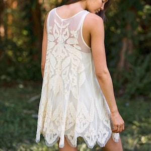 Puede incluir: Vestido sin mangas de color marfil con un panel trasero transparente y detalles de encaje floral intrincados. El vestido presenta una falda fluida y en capas y un dobladillo festoneado. La parte trasera del vestido tiene un dise&ntilde;o bordado delicado.