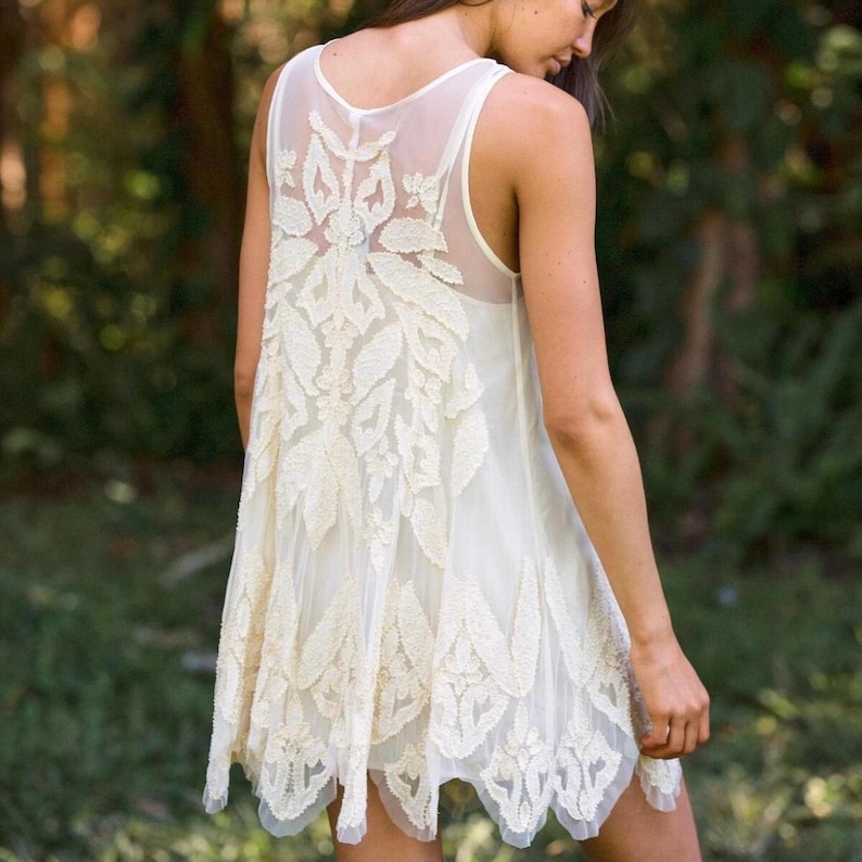 Puede incluir: Vestido sin mangas de color marfil con un panel trasero transparente y detalles de encaje floral intrincados. El vestido presenta una falda fluida y en capas y un dobladillo festoneado. La parte trasera del vestido tiene un dise&ntilde;o bordado delicado.