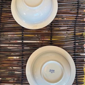 Op de afbeelding: Twee witte kommen en schotels van het merk Alessi worden getoond. De kommen staan ondersteboven en tonen het logo van het merk op de bodem. De kommen en schotels liggen op een bruine geweven mat met een bloemenpatroon.