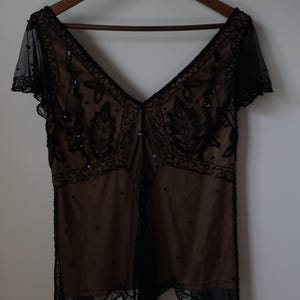 May include: A vintage-style black beaded top with a deep V-neck and short sleeves. The top features intricate beadwork and a sheer overlay, creating a delicate and elegant look. The garment is hanging on a wooden hanger.