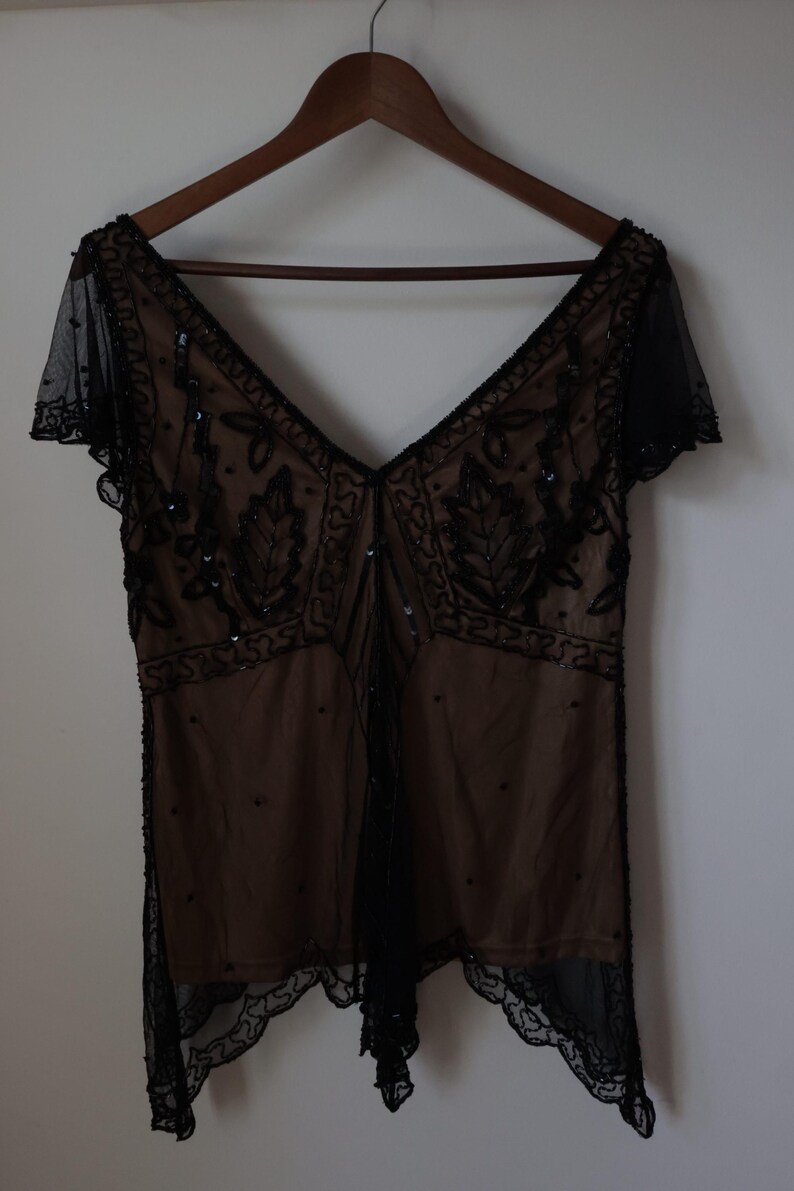 May include: A vintage-style black beaded top with a deep V-neck and short sleeves. The top features intricate beadwork and a sheer overlay, creating a delicate and elegant look. The garment is hanging on a wooden hanger.