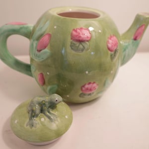 Puede incluir: Una tetera de cerámica verde, adornada con flores de loto rosas y lunares blancos. La tapa presenta una figura de rana. La tetera tiene un asa y un pico curvos, perfectos para servir té.