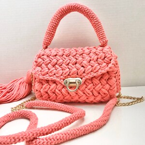 May include: A coral pink crocheted handbag with a gold chain strap and a tassel. The bag has a top handle and a flap closure with a gold clasp.