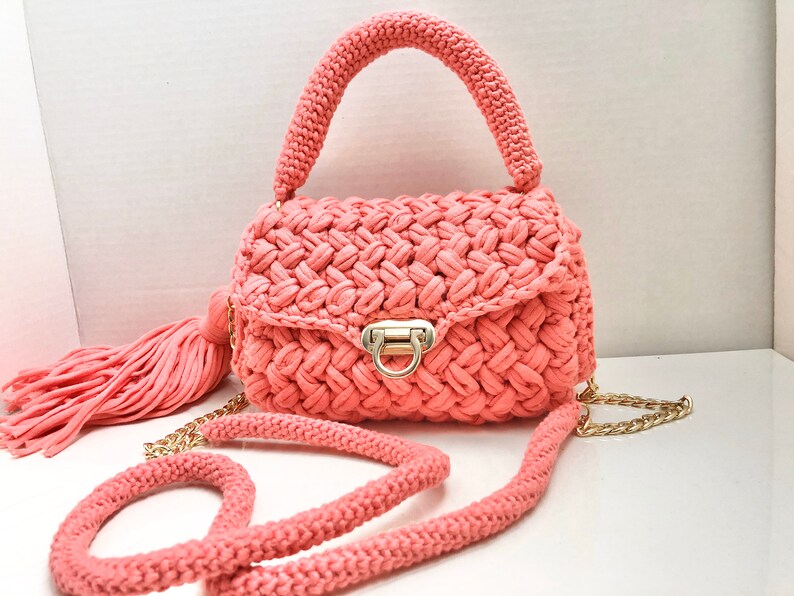 May include: A coral pink crocheted handbag with a gold chain strap and a tassel. The bag has a top handle and a flap closure with a gold clasp.