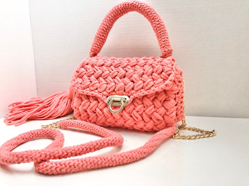May include: A coral pink crocheted handbag with a gold chain strap and a tassel. The bag has a flap closure with a gold clasp.