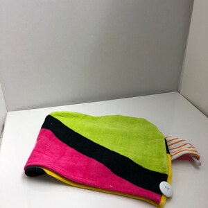 May include: A colorful hair towel with a button closure. The towel is green, black, pink, and yellow. It has a striped orange and white fabric tab.