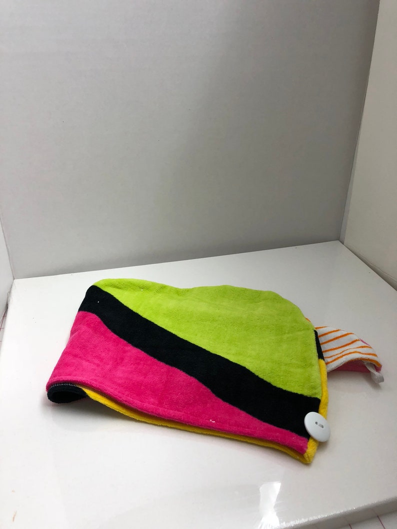 May include: A colorful hair towel with a button closure. The towel is green, black, pink, and yellow. It has a striped orange and white fabric tab.
