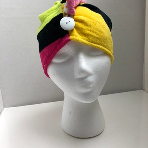 May include: A colorful headband with a black, yellow, pink, and green striped pattern. The headband has a white button and a pink and orange bow on top.