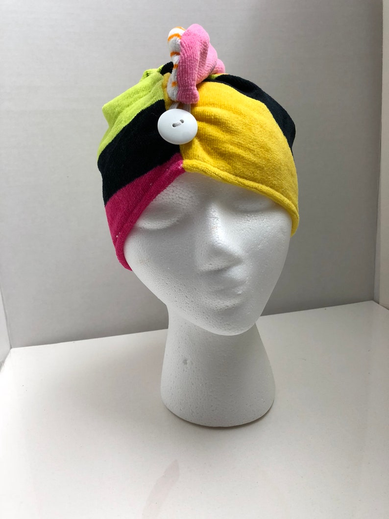 May include: A colorful headband with a black, yellow, pink, and green striped pattern. The headband has a white button and a pink and orange bow on top.