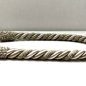May include: A pair of twisted rope curtain tiebacks in a light beige and white color. The tiebacks are made of a soft, woven fabric and have a tassel at each end.