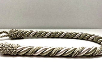 May include: A pair of twisted rope curtain tiebacks in a light beige and white color. The tiebacks are made of a soft, woven fabric and have a tassel at each end.