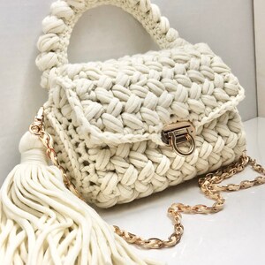 May include: A white crocheted handbag with a gold chain strap and a white tassel. The bag has a top handle and a flap closure with a gold clasp.