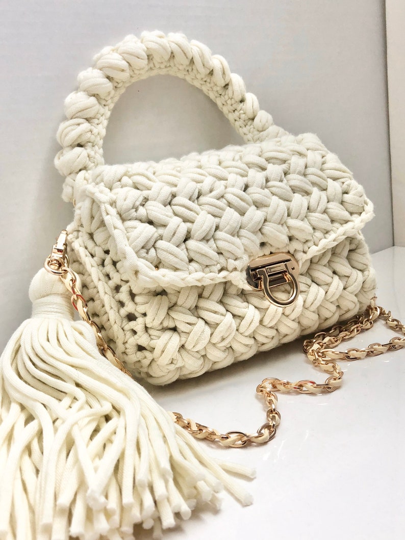 May include: A white crocheted handbag with a gold chain strap and a white tassel. The bag has a top handle and a flap closure with a gold clasp.